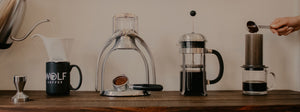 Water being poured from a kettle into a pour over coffee maker into a Wolf Coffee Mug, a manual espresso machine, a french press half full of coffee, and an aeropress being filled with coffee grounds from a silver scoop.