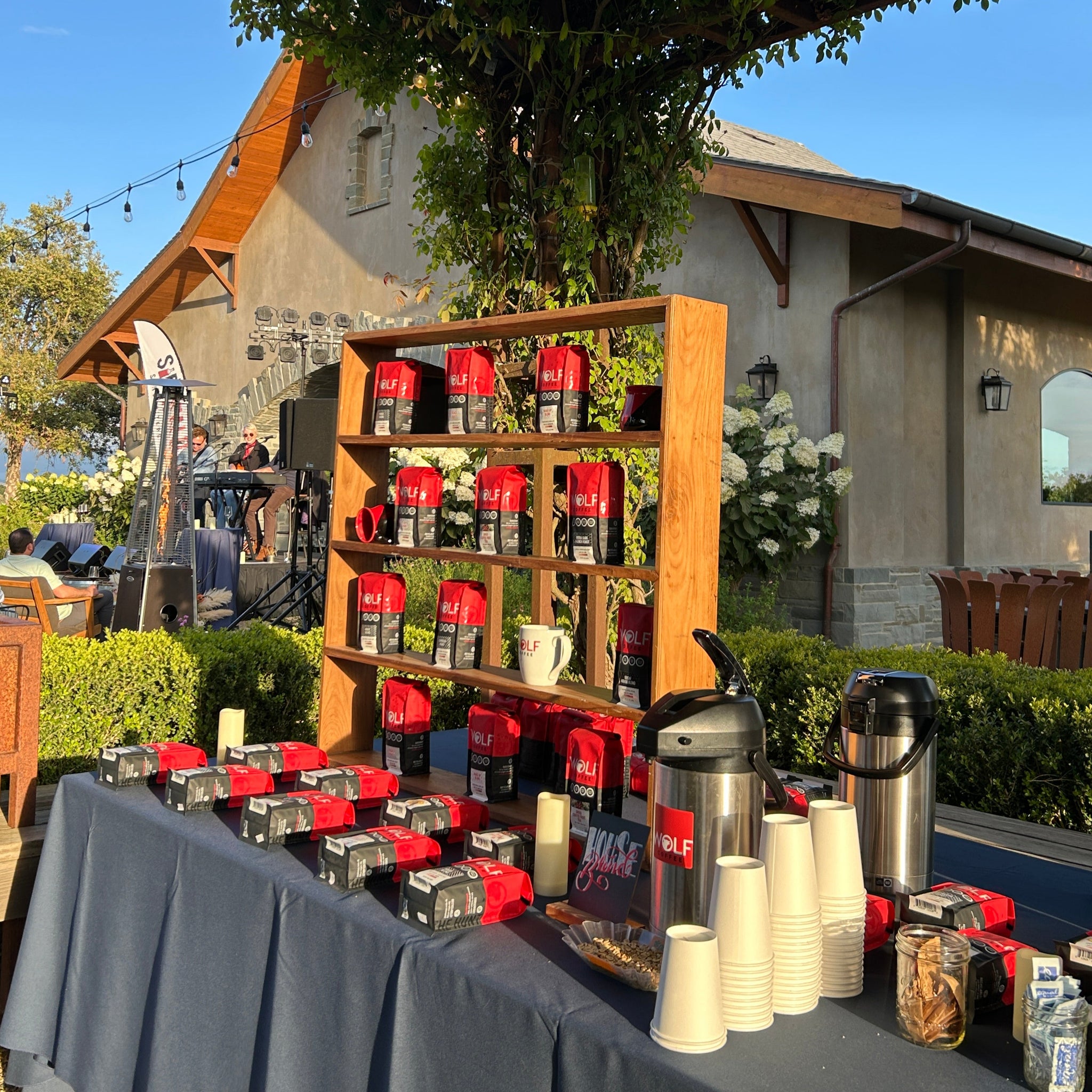 Outdoor setting with a wooden shelf displaying red and white bags of coffee, trees, and a clear blue sky.